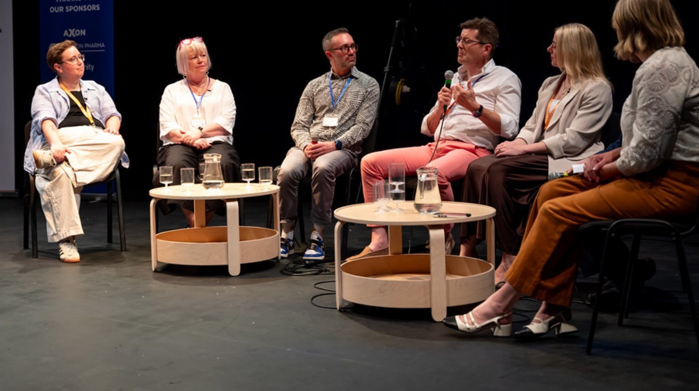 Dominic speaking into a microphone on a six-person panel on stage