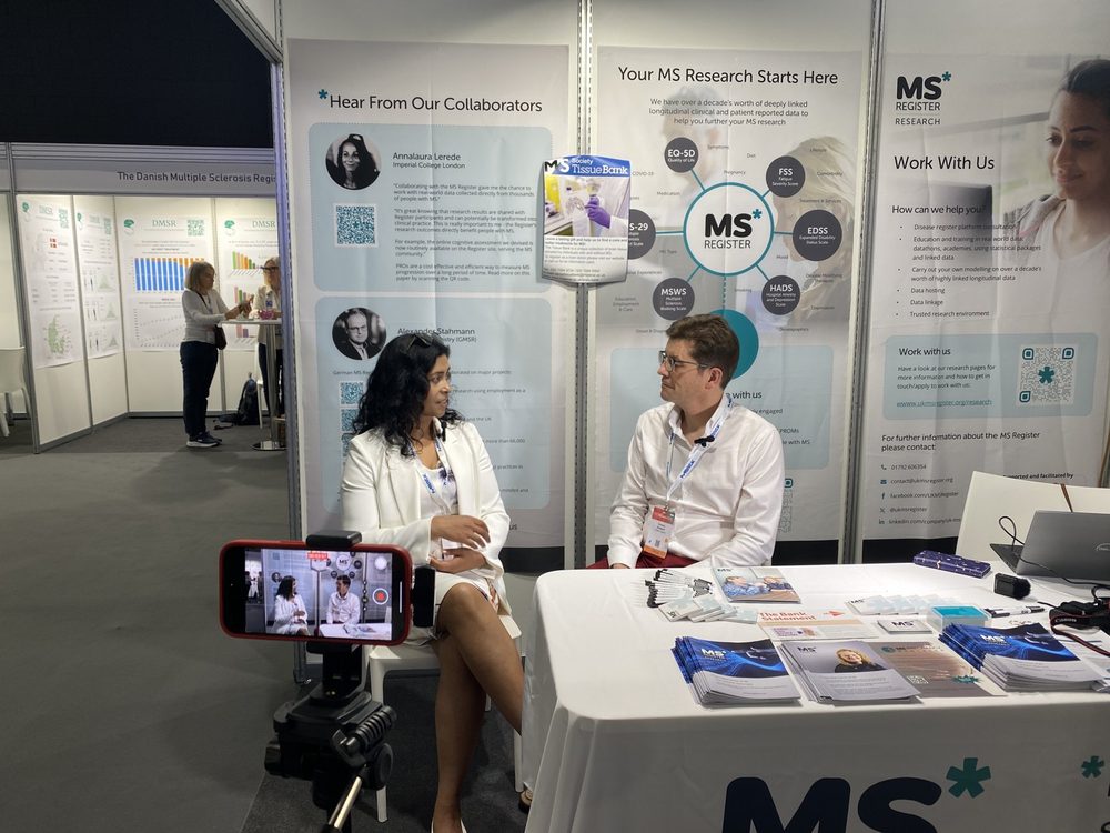 Dominic seated at a congress stand interviewing Dr Sharmilee Gnanapavan, with a phone on a tripod recording
